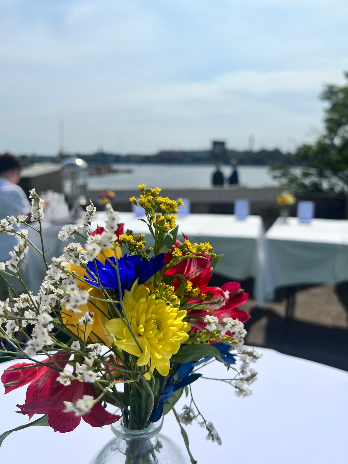 Färgglada sommarblommor i vas på vit duk, med vattnet i bakgrunden